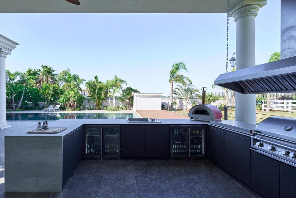 U-shaped Capo Cuòco outdoor kitchen on a covered porch: dark cabinet fronts under a stainless worktop, glass-front refrigeration along the back run, a dome pizza oven at the corner and a built-in gas grill beneath an extraction hood on the right-hand return