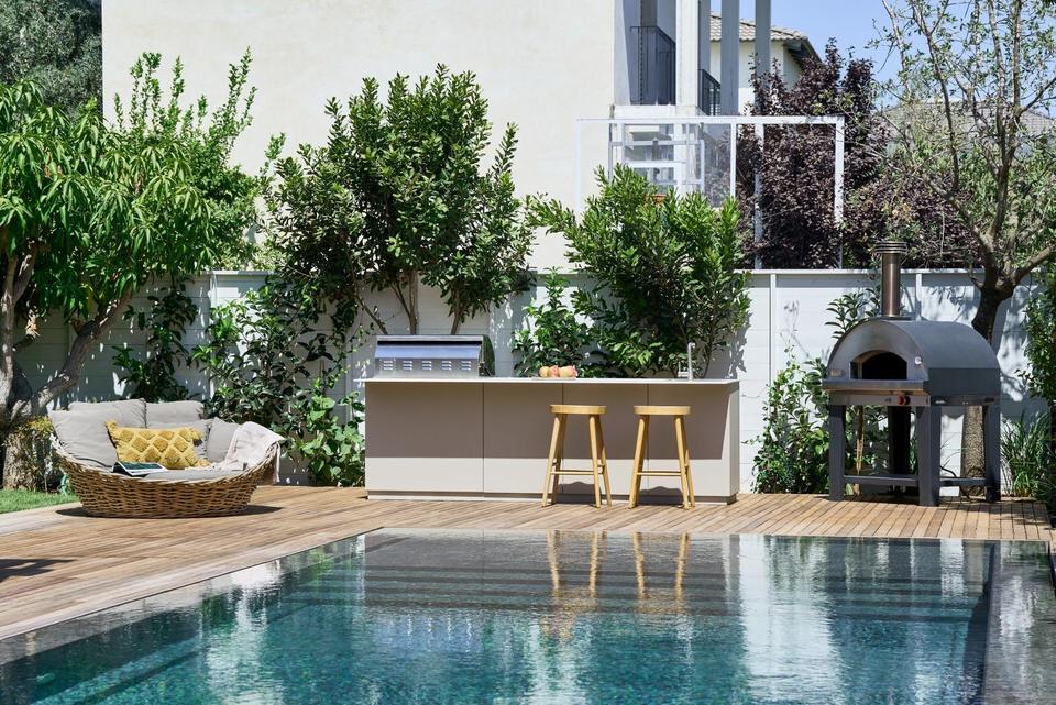 Pale UNICA outdoor kitchen island with a built-in gas grill and two bar stools, standing on a timber pool deck beside a swimming pool, with a freestanding pizza oven on a steel stand alongside